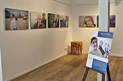 Der Ausstellungsraum mit den Fotos. Ein Aufsteller mit der Titeltafel lehnt an einer Säule in der Mitte des Raums.