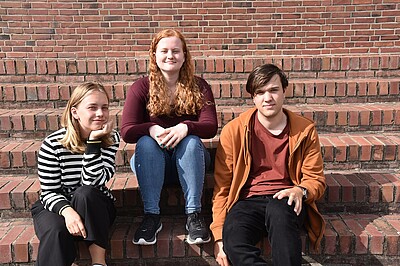 Drei junge Menschen sitzen auf einer Außentreppe und blicken in die Kamera. 