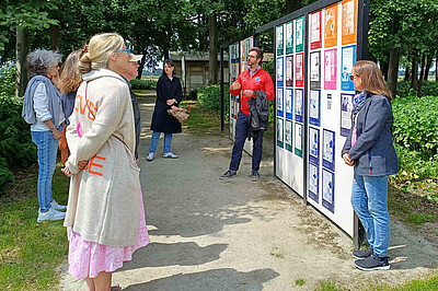 Mehrere Workshop-Teilnehmende stehen vor der Plakatwand des Ort der Verbundenheit während eine Person vom Projektteam zu ihnen spricht.