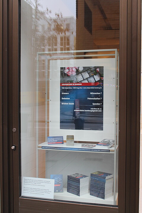 Ansicht des sogenannten Projekt-Fensters des Geschichtsort Stadthaus. Die Gestaltung wurde von der Stolperstein Initiative Hamburg übernommen. Ein Plakat, ein Stolperstein sowie auf der unteren Ebene des Ausstellungsmoduls sind die Publikationen gestapelt