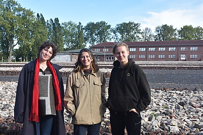 Three young women pose for a photo. Behind them you can see the stone formations which represent the former barracks and a brick building.