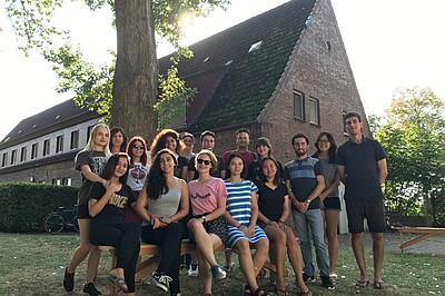 Junge Menschen sitzen und stehen als Gruppe und lächeln in die Kamera. Im Hintergund ein Haus und ein Baum.