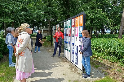 Menschen stehen vor einer Wand mit Postern, eine Person erklärt etwas, im Hintergrund Bäume.