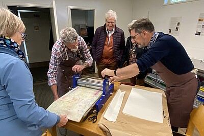 4 Personen sind um einen Tisch versammelt auf dem eine Druckerpresse steht, mehrere tragen Schürzen.