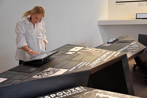 Eine Frau blickt interessiert auf einen Bildschirm, der auf der Ausstellungsplatte befestigt ist, bei der es um Zwangsarbeit geht.