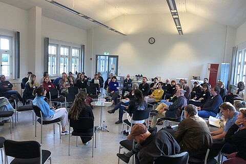 n der Mitte eines großen Raumes stehen um einen kleinen Tisch fünf Stühle im Kreis, ein Stuhl davon ist leer. Drum herum sind in mehreren Kreisen noch um die 60 Stühle, wo Teilnehmende der Tagung sitzen und zuhören.