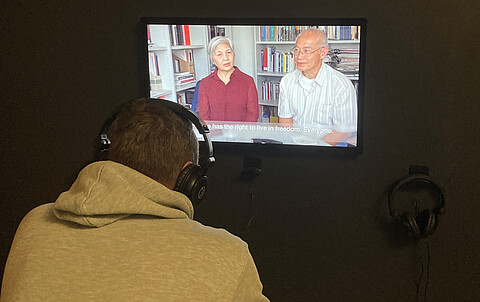 Videostation in der Ausstellung "Rechte Gewalt in Hamburg nach 1945" im St. Pauli Museum. Ein Mann im Hoodie sitzt vor einem Bildschirm mit Kopfhörern auf. Auf dem Bildschirm zwei Personen.
