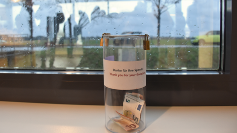 [Translate to English:] Eine Spendenbox aus Plastik, darin Geldscheine. Aufkleber "Danke für Ihre Spende". Im Hintergrund ein Fenster mit Regentropfen.