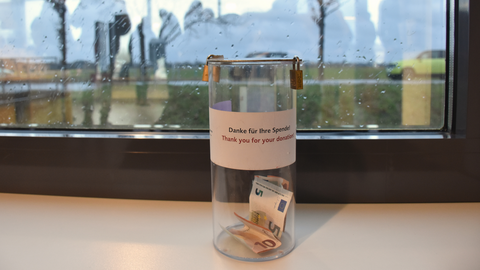 Eine Spendenbox aus Plastik, darin Geldscheine. Aufkleber "Danke für Ihre Spende". Im Hintergrund ein Fenster mit Regentropfen.
