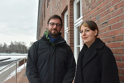 Ansgar und Natascha stehen draußen und blicken in die Kamera, hinter ihnen eine Treppe und eine Backsteingebäudewand. Es liegt Schnee.