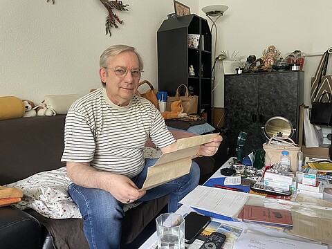  Harald Kröger sitzt in seinem Wohnzimmer. Er hat graue Haare und trägt eine Brille. Er hält ein historisches Dokument aus dem Nachlass in seiner Hand. 