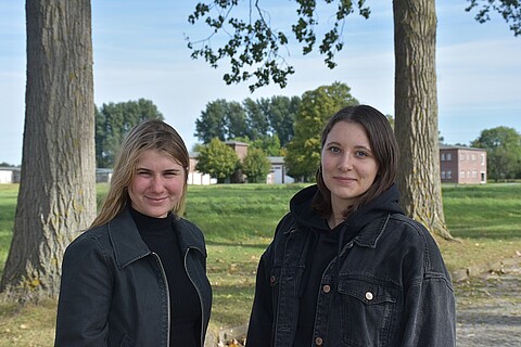 Zwei junge Frauen stehen nebeneinander und blicken in die Kamera, im Hintergrund das Außengelände der Gedenkstätte