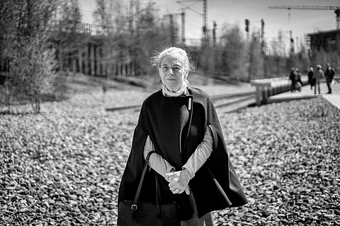 Eine weißhaarige Frau mit einem dunklen Cape und rotem Pullover steht am denk.mal Hannoverscher Bahnhof vor einem Steinfeld