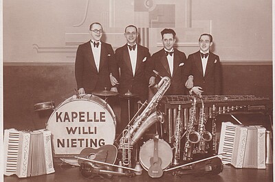 Schwarz-weiß Foto: Vier Männer in Anzügen mit Fliege, vor ihnen Instrumente, auf der Basstrommel steht Kapelle Willi Nieting