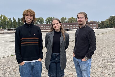 Es stehen die drei Freiwilligen Philipp, Uliana und Gabriel auf dem ehemaligen Appellplatz der KZ-Gedenkstätte Neuengamme