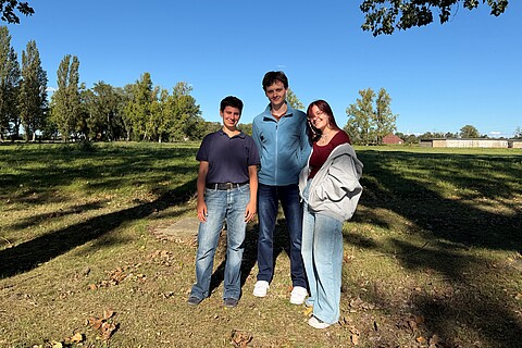Josefa, Jack und Arina stehen auf dem Gelände der Gedenkstätte vor einem Feld