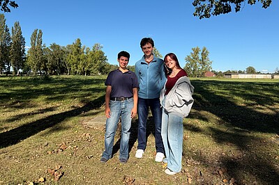 Josefa, Jack und Arina stehen auf dem Gelände der Gedenkstätte vor einem Feld