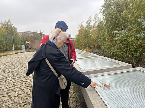 Eine ältere Frau und ein Mann stehen vor einer Namenstafel. Die Frau zeigt auf den unteren Abschnitt der Tafel. Hinter den Namenstafeln sind Bahngleise.