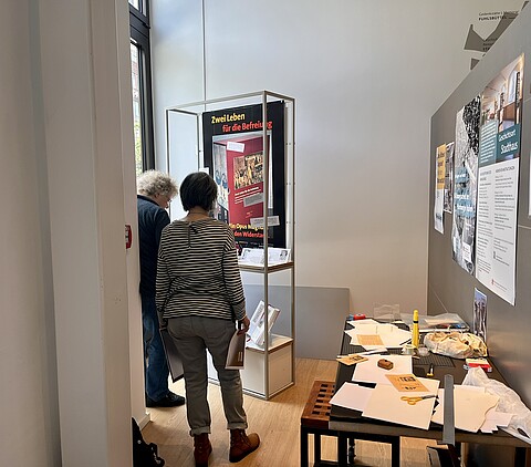 Zwei Personen arbeiten an der Ausstellungsgestaltung, im Vordergrund ein Tisch mit Papier und Materialien.