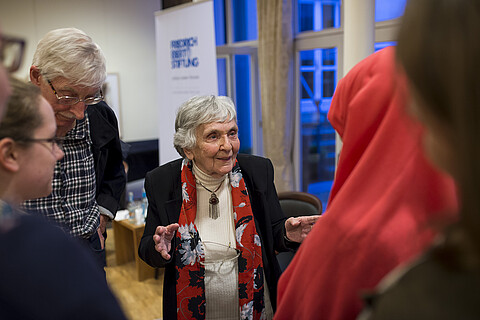 Eine alte Frau steht in der Mitte, ihr hören Menschen zu.