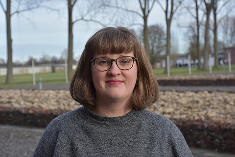 Clara Mansfeld trägt eine Brille und ein graues Kleid, sie blickt lächelnd in die Kamera, im Hintergrundbäume.