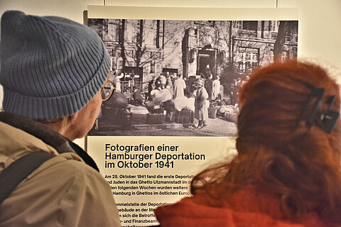 Zwei Besuchende stehen in der Ausstellung am Geschichtsort Stadthaus vor einer schwarzweiß Fotografie mit dem Text Fotografien einer Hamburger Deportation im Oktober 1941