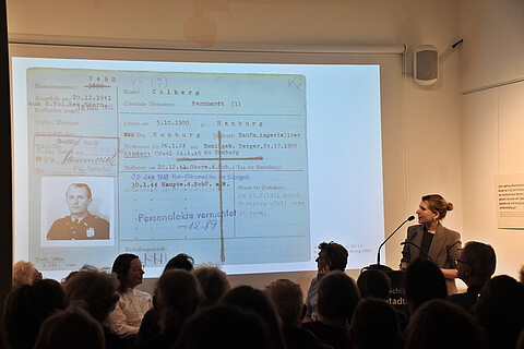 Johanna Schmied steht vor einer Leinwand mit einem alten Dokument mit einem Portraitfoto eines Polizisten, spricht über Colberg