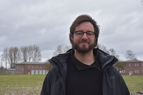 Ansgar Karnatz trägt eine Brille und eine schwarze Jacke. Ansgar lächelt in die Kamera, im Hintergund Wiese und Backsteingebäude.