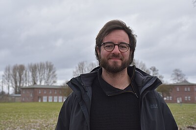 Ansgar Karnatz trägt eine Brille und eine schwarze Jacke. Ansgar lächelt in die Kamera, im Hintergund Wiese und Backsteingebäude.
