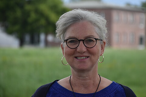 Dagmar Bonin trägt eine Brille und Ohrringe, sie blickt in die Kamera. Im Hintergund Wiese und Gebäude. 