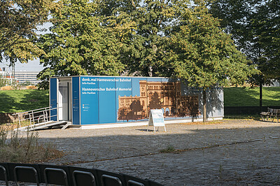 Container mit einer Rollstuhlrampe davor, beklebt mit dem Schrift und einem historischen Bild des Hannoverschen Bahnhofs, im Hintergrund Bäume, im Vordergrund ein Platz und Sitzgelegenheiten