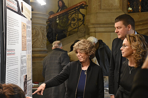 Drei Personen vor einer Ausstellungstafel, eine Person deutet auf ein Detail auf der Tafel