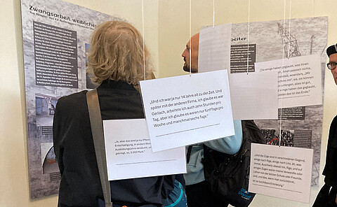 Blick in die Ausstellung "Mit Augen von heute", Personen stehen vor Tafeln, von der Decke hängen Zitate