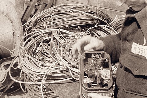 Eine schwarzweiß-Aufnahme, auf der viel Kabelgewirr zu sehen ist und im Vordergrund eine Hand, die ein einfaches selbstgebasteltes Radiogerät hält. Auf dem Ärmel des  Arms steht eine Nummer.