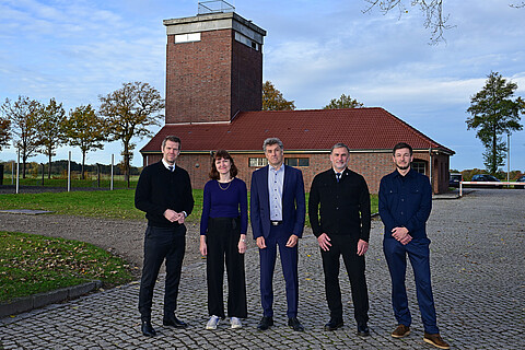 Fünf Personen in dunkelblauer Kleidung stehen am ehemaligen Wachturm in der KZ-Gedenkstätte Neuengamme