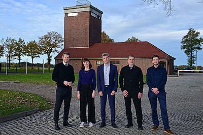 Fünf Personen in dunkelblauer Kleidung stehen am ehemaligen Wachturm in der KZ-Gedenkstätte Neuengamme