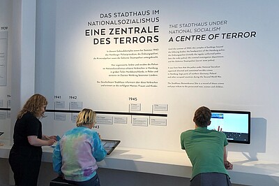 Drei Personen stehen vor einer Ausstellung und sehen sich etwas auf zwei Bildschirmen an. 