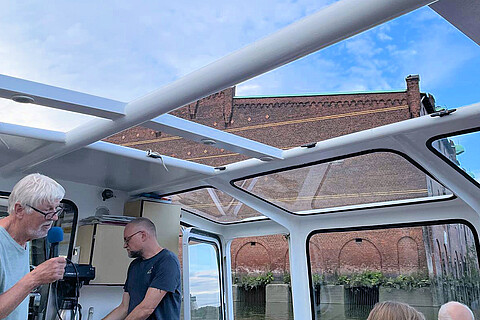 Hafenrundfahrt zum Dessauer Ufer mit einem Mann, der ins Mikrofon spricht. Durch die Barkassenfenster sichtbar ein Ziegelbau.