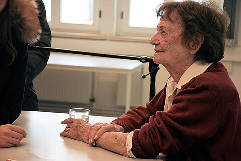 Dita Kraus sitzt an einem Tisch und hält ein Glas Wasser in der Hand, sie schaut zu einer anderen Person, von der nur die Hände und ein Teil des Oberkörpers im Bild sind.