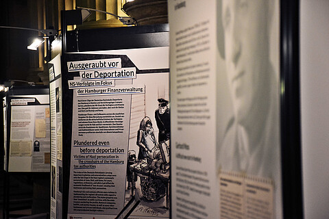 Blick in die Ausstellung "Ausgeraubt vor der Deportation" auf drei Tafeln. Eine Tafel trägt den Titel der Ausstellung, auf der vorderen Tafel ist ein Gesicht zu erkennen