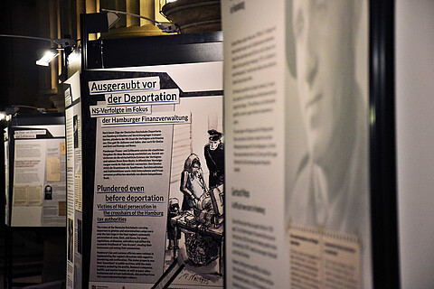 Blick in die Ausstellung "Ausgeraubt vor der Deportation" auf drei Tafeln. Eine Tafel trägt den Titel der Ausstellung, auf der vorderen Tafel ist ein Gesicht zu erkennen