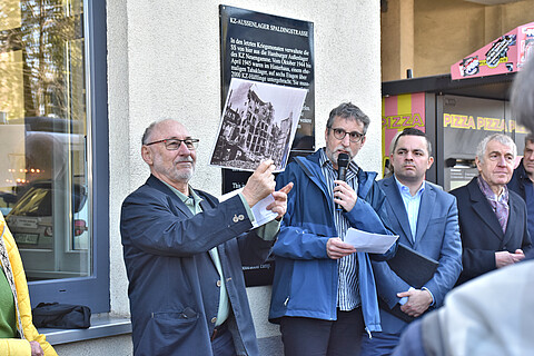 : Die Redner stehen vor dem Gebäude, einer hält ein historisches Foto in die Höhe, auf der das zerstörte Gebäude zu sehen ist, in dem 1944 das KZ-Außenlager eingerichtet wurde. 