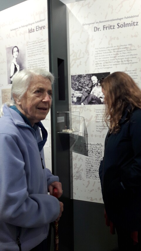 Brigitte Alexander steht in einer Ausstellung vor der Vitrine, die die Taschenuhr ihres Vaters zeigt, im Hintergrund eine Tafel mit einem schwarzweiß Foto von ihr und ihrem Vater, 2017