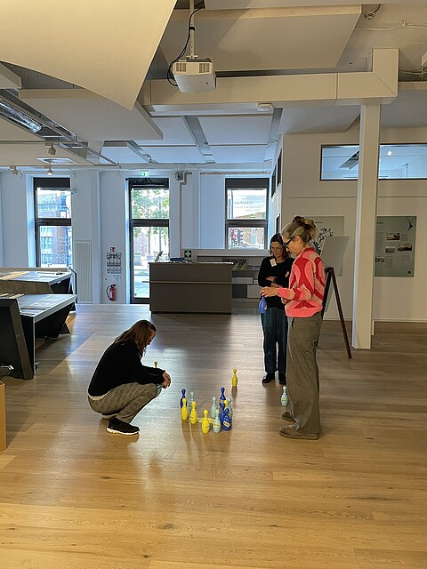 Drei Workshop-Teilnehmende, zwei stehend, eine kniend, setzen Plastikkegel in mehreren Farben auf den Boden für eine pädagogische Übung.