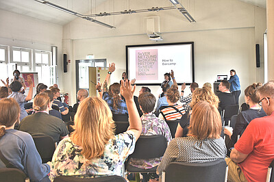 Personen sitzen auf Stühlen und schauen nach vorne, wo "How to Barcamp" auf einer Leinwand steht und 2 Personen mit Mikrofon in die Gruppe schauen