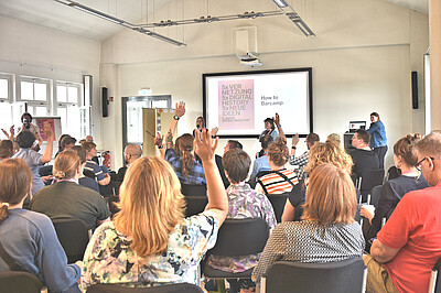 Personen sitzen auf Stühlen und schauen nach vorne, wo "How to Barcamp" auf einer Leinwand steht und 2 Personen mit Mikrofon in die Gruppe schauen