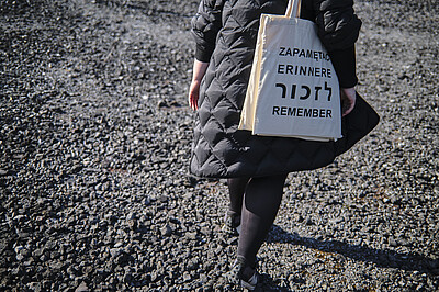 Person in schwarzer Leggings und mit schwarzem Mantel auf dunklem Schotter laufend. Der Rücken und die Beine sind Fotographiert, nicht der Kopf, sie trägt eine Tasche auf der in mehrerern Sprachen "Erinnern" steht.