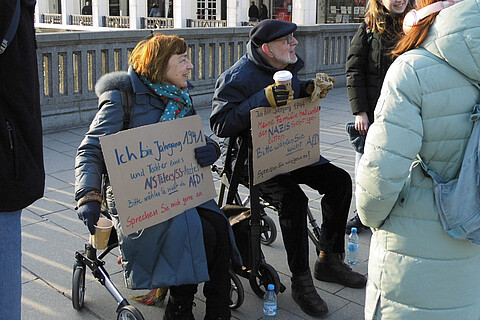 Zwei ältere Menschen sitzen auf Rollatoren und halten Plakate in der Hand. Um sie herum mehrere andere Personen. Im Hintergrund die Arkaden an der Alster.