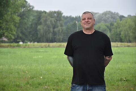 Andreas Strippel steht auf dem Gelände der KZ-Gedenkstätte Neuengamme. Im Hintergrund sind eine grüne Wiese und Bäume zu sehen. und 