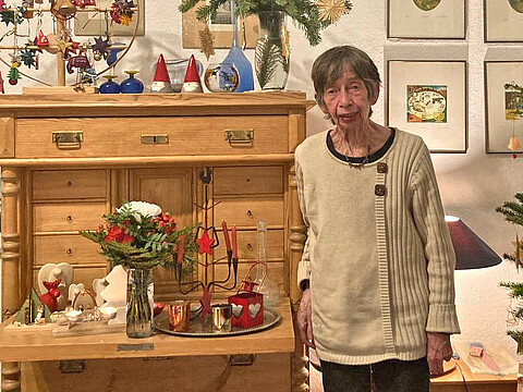 Eine ältere Dame steht vor einem Holzschrank mit Dekorationsobjekten